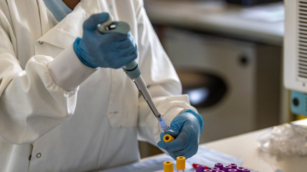 A person in a white lab coat and blue gloves uses a pipette to inject liquid into a vial.